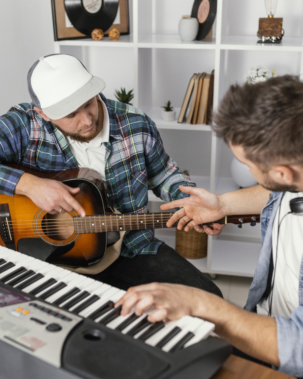 Two men are sitting indoors; one is playing an acoustic guitar while the other plays an electronic keyboard, much like music theory teachers near me guiding their students through melodies.