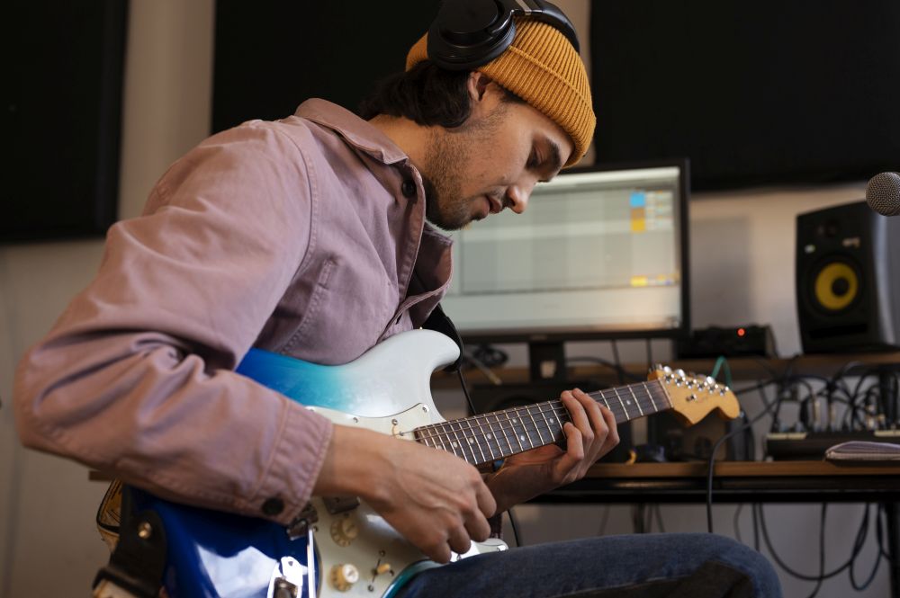 Person wearing headphones and a yellow beanie plays an electric guitar in a recording studio, exploring Electric Guitar Lessons Singapore to Play Your Favourite Songs with computer and audio equipment in the background.