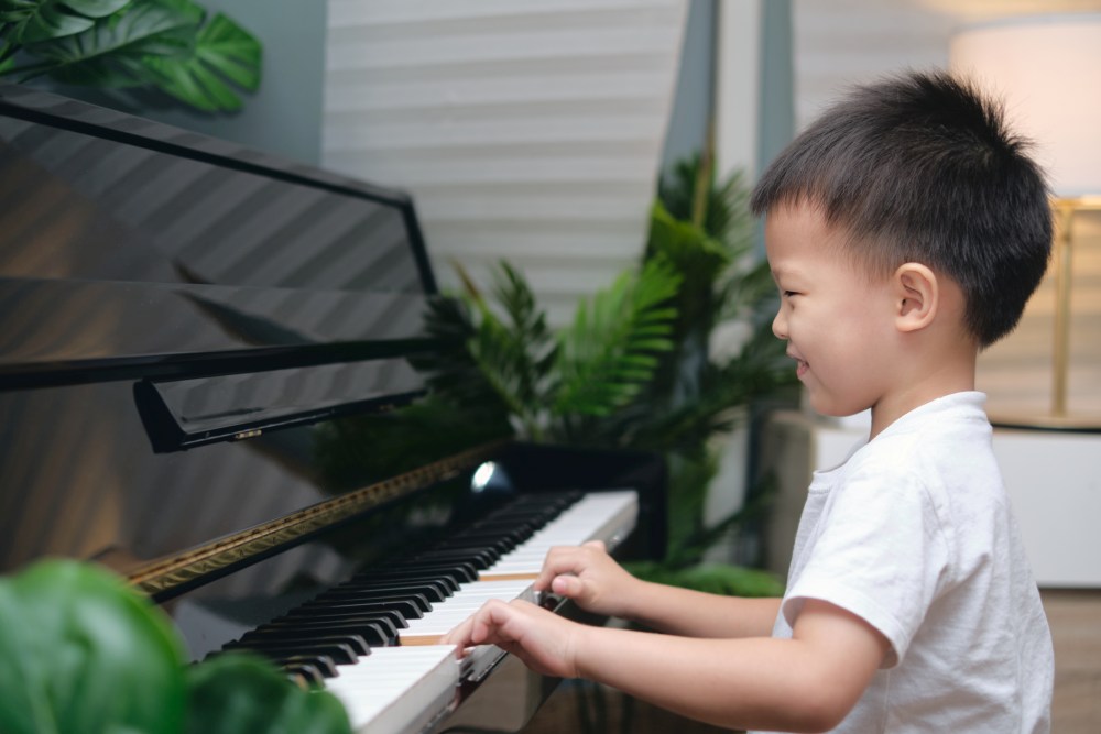 A young child in a white shirt plays the piano indoors, surrounded by green plants and soft lighting, enjoying a Piano Class for Kids Singapore in a fun and engaging environment.