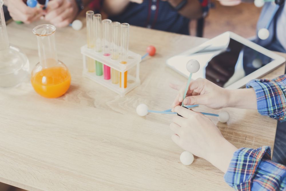 Hands assemble a molecular model on a table with test tubes, a flask containing orange liquid, and a tablet device in the background—illustrating concepts that can help boost PSLE Science Tuition: How to Strengthen Exam Readiness through interactive learning.